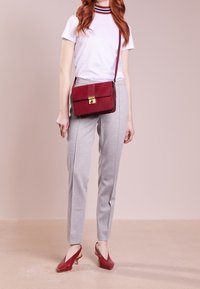 Röd lädercrossbody-väska med guldfärgad hårdvara, kombinerad med en vit T-shirt och grå skräddarsydda byxor. Modellen bär röda spetsiga klackar.
