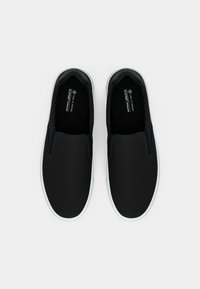 Chaussures noires à enfiler avec une tige lisse, une semelle en caoutchouc blanche et un marquage discret à l'intérieur. Conçues avec un matériau vegan pour la durabilité.