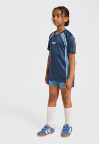 Marinblå sporttröja med ljusblå detaljer, kombinerad med matchande blå shorts. Bär blå skor med vita detaljer och cremefärgade knähöga strumpor.