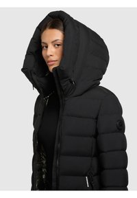 Kvinde iført en sort polstret jakke med stor hætte, halvt lynet op, mod en ensfarvet lys grå baggrund.