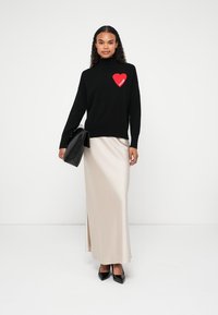 Woman wearing a black sweater with a red heart labeled "Future," a long beige skirt, black heels, and holding a black clutch bag.