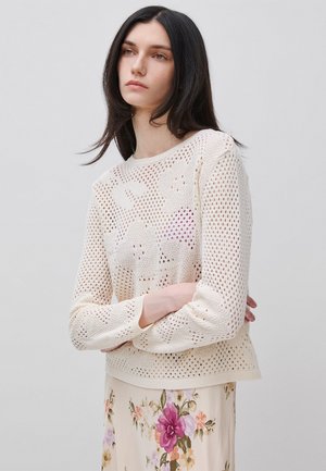 Donna dai lunghi capelli scuri indossa un maglione crema a maglia aperta sopra un vestito fiorato, in piedi davanti a uno sfondo grigio chiaro uniforme.