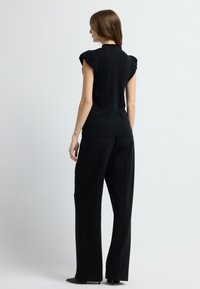 Zwart shirt met korte mouwen en gerimpelde schouders, gecombineerd met losse zwarte broek en puntige hakken, weergegeven vanuit een iets gedraaide achteraanzicht.