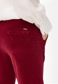 Rode corduroy broek met een gladde textuur, voorzien van een achterzak met een zilveren accent en een op maat gemaakte taille, gecombineerd met een witte blouse.