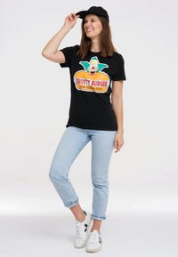 Black t-shirt featuring a colorful "Krusty Burger" graphic. The model wears light blue jeans and white sneakers with black accents.