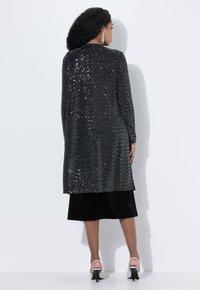 Larga chaqueta negra con lentejuelas y una textura suave, con un corte recto y espalda ligeramente más larga. La falda negra y los tacones plateados completan el conjunto.