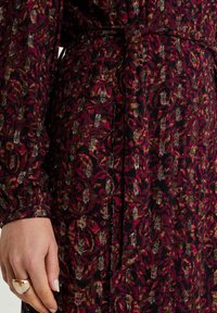 Vävd klänning i mörkröd och svart med blommönster och metalliska accenter. Den har långa ärmar och en bälte i midjan.