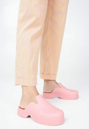 Pink rubber clogs with a rounded toe and open back, featuring a smooth texture. Worn with striped, ankle-length pants in pastel colors.