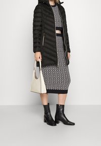 Schwarze Steppjacke mit diagonaler Steppung, kombiniert mit einem gemusterten schwarz-weißen Kleid. Schwarze Stiefel und eine kleine cremefarbene Handtasche.