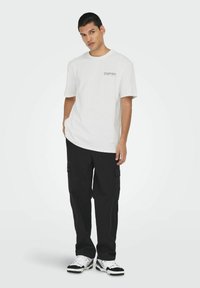 T-shirt blanc en coton pour homme avec un col rond et des manches courtes, associé à un pantalon cargo noir avec des poches latérales et une coupe décontractée.