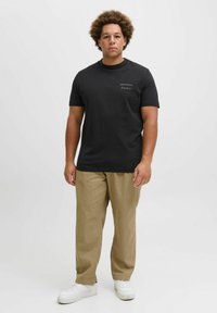 Sort bomulds t-shirt med korte ærmer, prydet med hvid tekst "ORIGINAL Studio" på venstre bryst, parret med beige bukser og hvide sneakers.