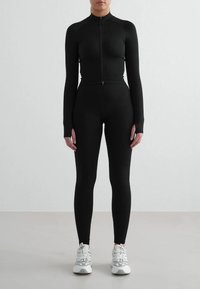 Haut noir à manches longues zippé avec un col montant, associé à des leggings noirs moulants. Le mannequin porte des baskets blanches avec des détails texturés.