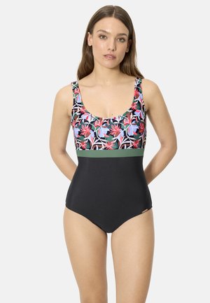 Mujer vistiendo un bañador de una pieza con la parte superior de estampado floral, la parte inferior negra sólida y una banda verde en la cintura, de pie contra un fondo claro.