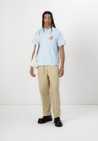 Light blue cotton T-shirt with graphic design, paired with beige pants and black chunky shoes. Model holds a white tote bag.
