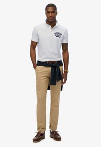 Camiseta polo blanca con logotipo marino, chinos beige y zapatos marrones. Un suéter marino está atado a la cintura, con tejido suave y diseño clásico.