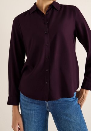 Mujer vestida con camisa de manga larga abotonada de color púrpura oscuro combinada con jeans azules, de pie con una mano apoyada en la cadera.