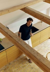 Jeune homme aux cheveux bouclés, portant une chemise noire, un pantalon blanc et des sandales, appuyé contre un comptoir de cuisine en bois avec un évier et un bol de fruits visibles.