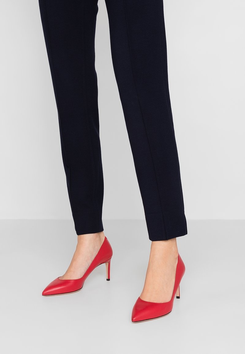 Femme portant un pantalon noir ajusté et des escarpins rouges à bout pointu sur un fond uni clair.