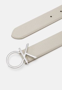 Cintura in pelle beige chiaro con superficie testurizzata, dotata di una fibbia in metallo argentato a forma di "CK" all'interno di un cerchio.