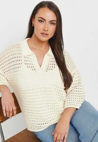 Krämfärgad stickad tröja med V-hals, korta ärmar och öppen, prickig mönstring. Den är ihop med ljusblå jeans och sitter på en trästol.