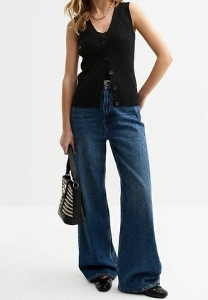 Femme portant un haut noir sans manches à boutons, un jean bleu à jambes larges, des chaussures noires, un collier chaîne en or, et tenant un sac à main noir à motifs.