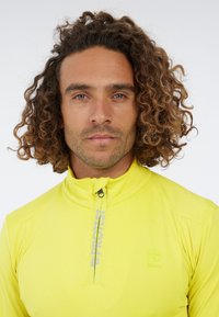 Man met krullend haar en een lichte baard, gekleed in een felgele pullover met kwartrits en een 'Protest'-logo op de borst, kijkt recht vooruit.