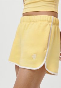 Pantaloni corti gialli realizzati in un tessuto morbido con cintura regolabile, bordo bianco e logo ricamato sulla parte bassa della gamba sinistra.