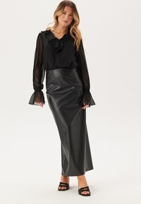 Svart blus med volangkrage och genomskinliga ärmar, tillsammans med en maxikjol i svart konstläder. Modellen bär svarta sandaler.