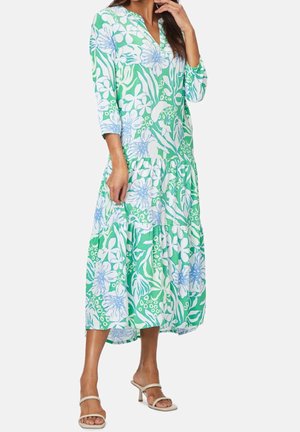 Mujer con vestido midi floral en verde, blanco y azul, con mangas 3/4 y escote en V, combinada con sandalias beige de tacón, de pie frente a un fondo liso.