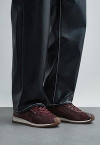 Crne široke hlače uparene s bordo teksturiranim tenisicama oznake Michael Kors stoje na sivoj podlozi.