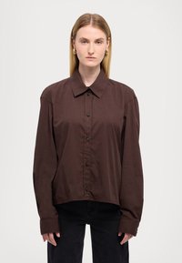 Camisa marrón de botones con cuello estructurado y mangas largas, con un corte relajado y una textura sutil. Combina con pantalones negros.