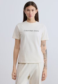 Κρεμ βαμβακερό μπλουζάκι με κοντά μανίκια, στρογγυλή λαιμόκοψη και μαύρο κείμενο "Calvin Klein Jeans" τυπωμένο στο κέντρο. Λεία υφή, καθημερινό σχέδιο.