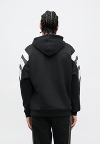 Čierna mikina s kapucňou s pruhovanými bielymi a sivými akcentmi na rukávoch. Mäkká textúra, voľný strih, žebrované manžety a lem. Vhodná na bežné nosenie.