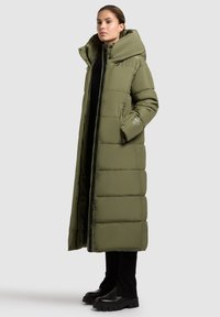 Garšs olivu zaļš pūkains mētelis ar augstu apkakli, ar rāvējslēdzēju priekšā, sānu kabatām un kvalitāti satura dizainu. Apvienots ar melniem zābakiem.