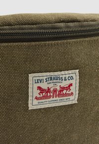 Close-up van een olijfgroene stoffen tas met het label van Levi Strauss & Co, waarin twee rode paarden een spijkerbroek uit elkaar trekken.