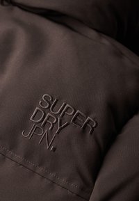 Chaqueta de tela marrón con bordado del logo "SUPER DRY JPN.", que muestra una textura suave con costuras visibles y líneas limpias.