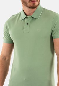 Polo verde hecho de algodón texturizado, con un cuello, dos botones marrones y un logotipo bordado en el lado izquierdo del pecho.