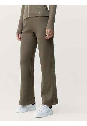 Personne portant un pantalon large côtelé vert olive, une veste zippée assortie et des baskets blanches avec des détails noirs au talon, debout sur un fond uni.