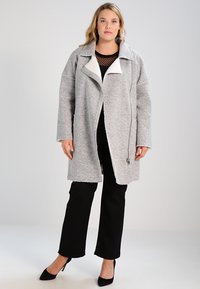 Abrigo texturizado gris con cuello blanco, de corte oversize, cierre frontal con cremallera y dos bolsillos laterales, combinado con pantalones negros y tacones.