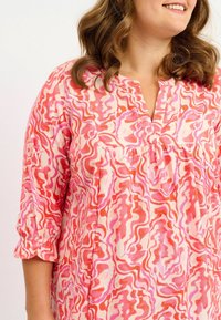 Fließende, rosa gemusterte Bluse mit kurzen Ärmeln, die abstrakte, gewellte Designs in Rot-, Rosa- und Cremetönen zeigt. Mit Knopfleiste am Ausschnitt.