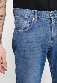 Jeans de ganga azul com corte reto, bainha desgastada, rebites de cobre e costura detalhada nos bolsos. Mão repousando ao lado.