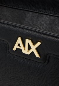 Bolso de mano de cuero texturizado negro que presenta un prominente acento en oro con el logo "AX". Las costuras añaden un interés visual al diseño.