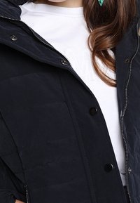 Tmavě modrá prošívaná bunda s kapucí, zipem a druky. Má prošitou texturu a je nošena přes bílé tričko s raženým nápisem.