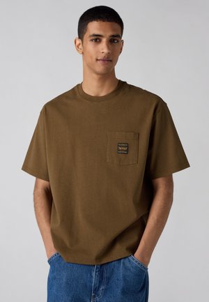 Joven con pelo corto y negro, vestido con una camiseta marrón oliva con un pequeño bolsillo en el pecho y vaqueros azules, con las manos en los bolsillos, sobre un fondo gris.