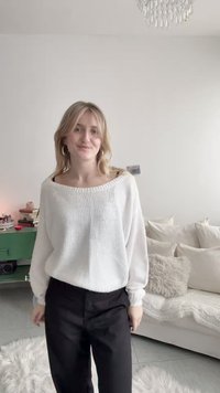 Νεαρή γυναίκα με ξανθά μαλλιά, που φοράει λευκό πουλόβερ και μαύρο παντελόνι, στέκεται σε ένα ζεστό σαλόνι με καναπέ και πράσινη ντουλάπα.