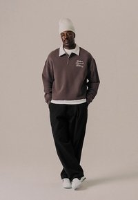 Uomo che indossa un felpa marrone con colletto bianco, pantaloni neri, sneakers bianche e un berretto di maglia bianco, in piedi con le mani in tasca contro uno sfondo neutro.