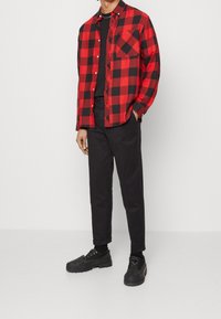 Camisa de cuadros rojo y negro con botones y un bolsillo en el pecho, combinada con pantalones chinos negros y zapatos negros con cordones. Textura suave, diseño casual.