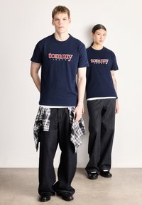 Camisetas de algodón azul marino con el logo "tommy JEANS", combinadas con jeans oscuros y holgados de mezclilla. Camisa de cuadros atada alrededor de la cintura, zapatos negros.