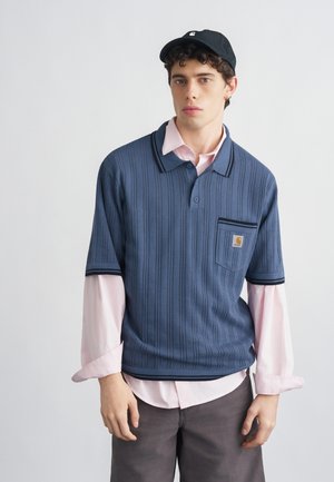 Mladý muž v čiernej čiapke, modrej krátkoročnej pletenej polokošeli cez svetloružovú košeľu s dlhým rukávom a tmavých nohaviciach, stojaci pred jednoduchým pozadím.