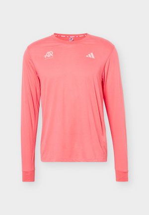 Mann mit dunklen Haaren, der ein korallenfarbenes Langarmshirt mit weißem grafischem Text und Symbolen auf der Rückseite trägt, kombiniert mit marineblauen Hosen.